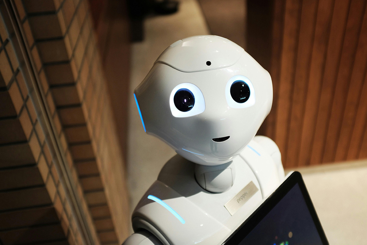 Humanoider Roboter mit großen runden Augen und weißem Gehäuse blickt in die Kamera, während er ein Tablet hält - symbolisiert den Einsatz moderner Technologien in Lehr- und Lernumgebungen.