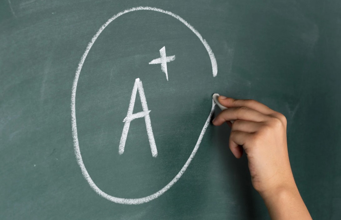 Hand mit Kreide schreibt ein 'A+' in einen Kreis auf eine grüne Tafel - symbolisiert Leistungsbewertung und das Streben nach Exzellenz im Kontext von KI-gestützten Prüfungsmethoden.