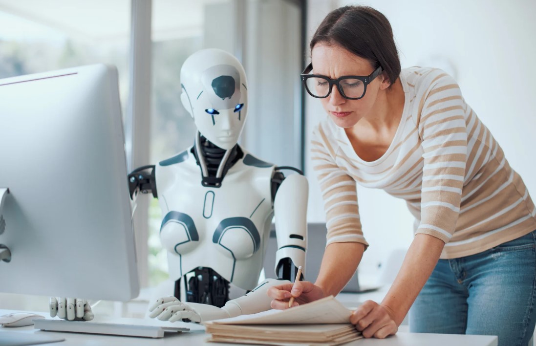 Eine Frau mit Brille arbeitet am Schreibtisch neben einem weißen humanoiden Roboter, beide blicken auf Dokumente - symbolisiert die Zusammenarbeit zwischen Mensch und künstlicher Intelligenz als didaktische Unterstützung in der Lehre.