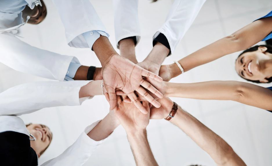 Mehrere Hände verschiedener Personen werden im Kreis zusammengelegt, Blick von unten nach oben - symbolisiert Teamwork, Zusammenhalt und erfolgreiche Gruppenarbeit in Lehrveranstaltungen.