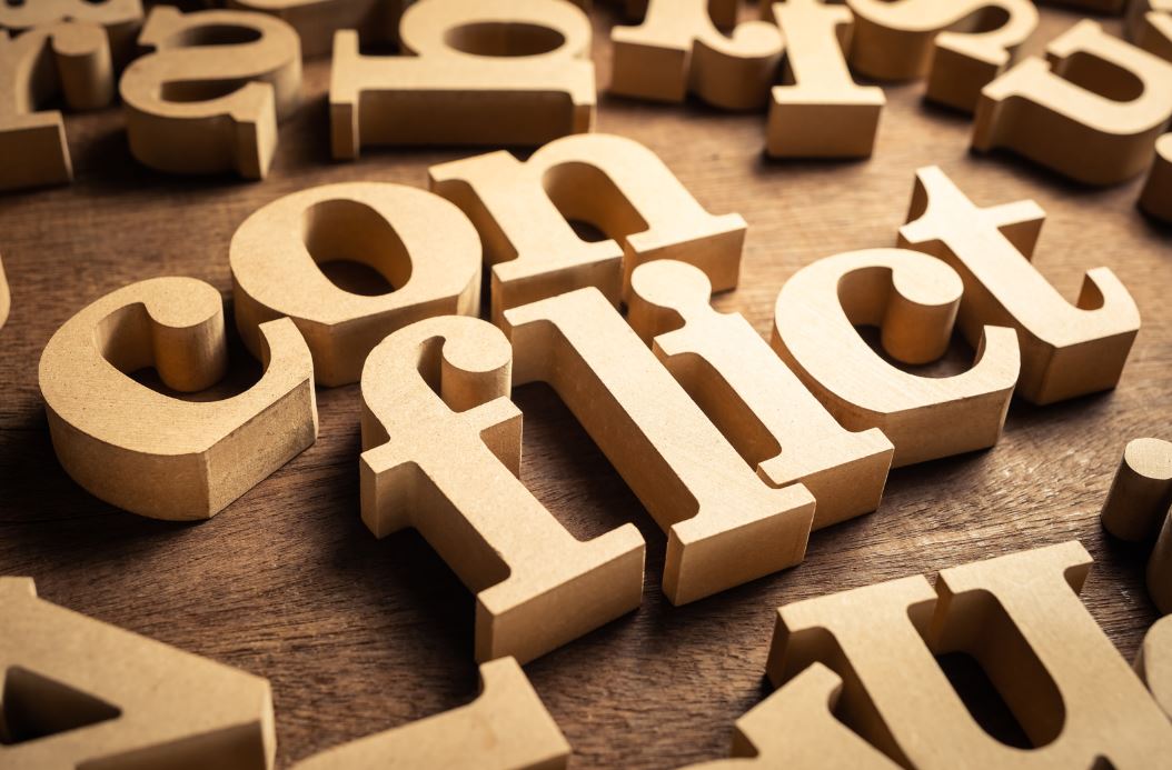 Holzbuchstaben, die das Wort 'conflict' bilden, umgeben von weiteren verstreuten Buchstaben auf Holzuntergrund - symbolisiert die strukturierte Auseinandersetzung mit Konflikten und deren Lösung in der Hochschullehre