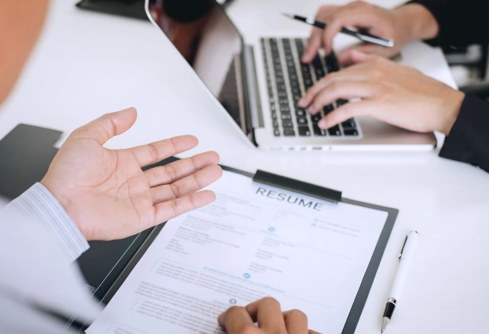 Person im Gespräch gestikuliert mit ausgestreckter Hand, während im Hintergrund ein Lebenslauf auf dem Tisch liegt und eine andere Person am Laptop arbeitet - symbolisiert professionellen Austausch, Beobachtung und gegenseitiges Lernen in der kollegialen Hospitation.