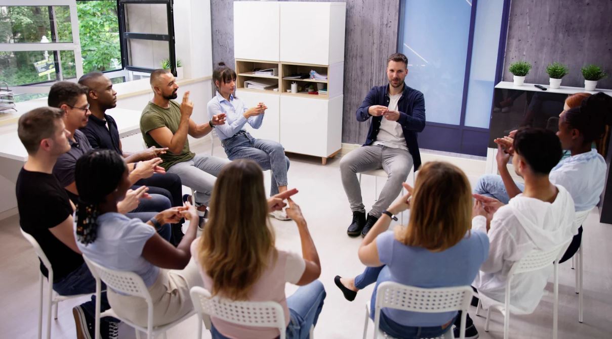 Gruppe von Menschen sitzt im Kreis in einem modernen Raum, während eine Person in der Mitte stehend spricht und gestikuliert - symbolisiert rhetorische Fähigkeiten, Präsentationstechniken und den Aufbau von Sprechkompetenz in der Hochschullehre.