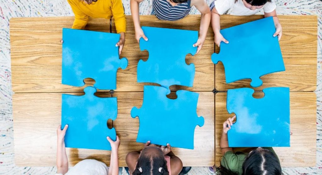 KI Post Card Sechs Personen sitzen um einen Holztisch und halten jeweils große blaue Puzzleteile in den Händen, Blick von oben - symbolisiert Zusammenarbeit, das Zusammenfügen verschiedener Perspektiven und die Dynamik von Gruppenprozessen.