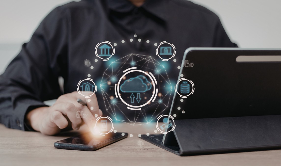 Person am Laptop mit Smartphone, über dem holografische Cloud- und Netzwerk-Symbole schweben, umgeben von kreisförmigen Icons für verschiedene digitale Dienste - symbolisiert die Vernetzung und Vielfalt von KI-Tools und digitalen Workflows in der Hochschullehre.